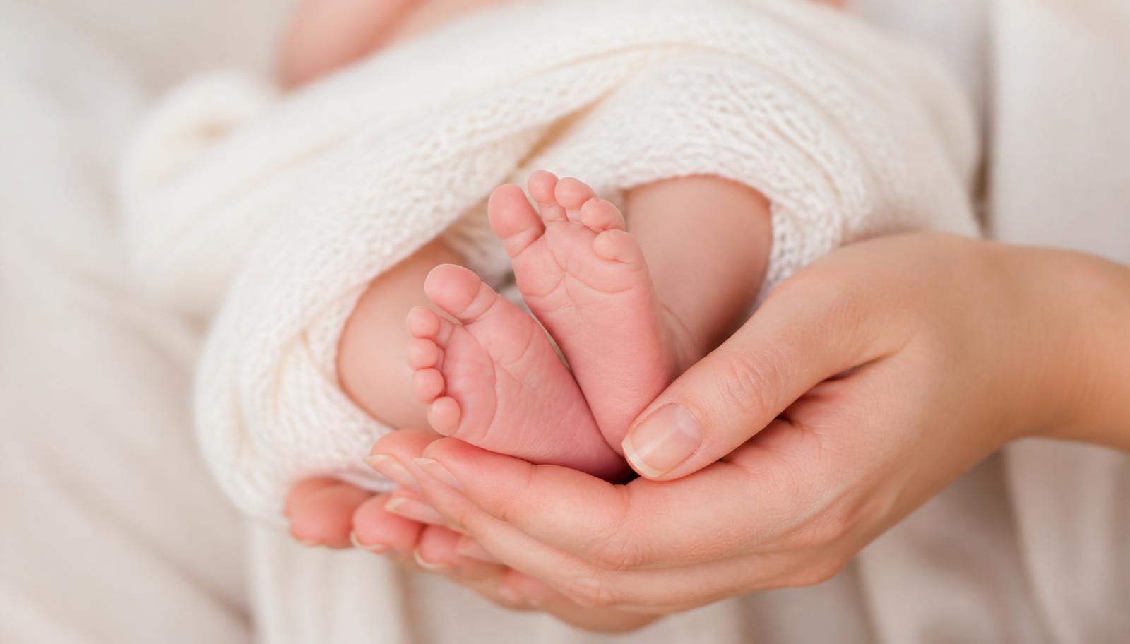 Parent's hands gently holding a baby's tiny feet wrapped in a cream knitted blanket in warm window light